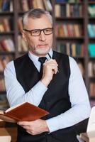 à la recherche d'inspiration. homme senior aux cheveux gris réfléchi en tenues de soirée tenant un bloc-notes et regardant ailleurs tout en se penchant à la table et avec une étagère en arrière-plan photo