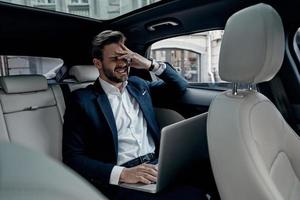 jeune homme frustré en costume complet travaillant à l'aide d'un ordinateur portable et couvrant le visage avec la main tout en étant assis dans la voiture photo