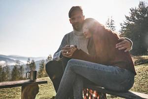 il est toujours là pour elle. beau jeune couple prenant un café le matin assis sur le banc dans les montagnes photo