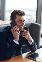 souriant jeune homme d'affaires en costume complet parlant sur le téléphone intelligent tout en étant assis dans le restaurant photo