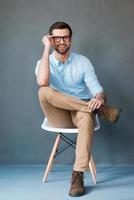 style créatif. toute la longueur du beau jeune homme ajustant ses lunettes et souriant à la caméra tout en étant assis sur fond gris photo