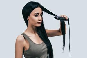 il est temps de faire une nouvelle coiffure séduisante jeune femme en tenue décontractée coiffant ses longs cheveux avec un fer à friser en se tenant debout sur fond gris photo