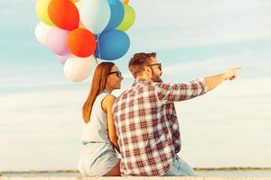 cette vue est magnifique jeune couple souriant regardant loin assis sur un parapet avec des ballons colorés photo