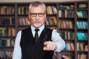 je vous choisis un homme senior confiant aux cheveux gris en tenue de soirée vous pointant du doigt tout en se tenant contre une étagère photo