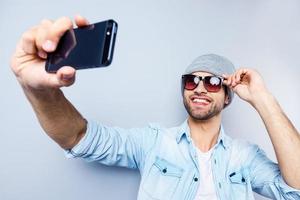 selfie vue de dessus du beau jeune homme au chapeau et lunettes de soleil faisant selfie et souriant debout sur fond gris photo