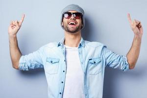 regardez ce beau jeune homme élégant en lunettes de soleil et chapeau pointant vers le haut et souriant en se tenant debout sur fond gris photo