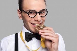 je suis un jeune homme joyeux gagnant en noeud papillon tenant une médaille d'or et la mordant debout sur fond gris photo