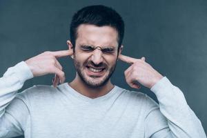 son trop fort frustré jeune homme exprimant la négativité tout en couvrant les oreilles avec les mains et debout sur fond gris photo