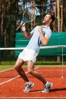 célébrer le succès. vue latérale d'un jeune homme heureux en polo tenant une raquette de tennis et faisant des gestes en se tenant debout sur un court de tennis photo