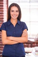 expert jeune et talentueux. jolie jeune femme gardant les bras croisés et souriant tout en se penchant sur son lieu de travail photo