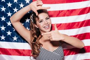concentrez-vous sur moi jeune femme joyeuse faisant un cadre de doigt et regardant à travers avec le sourire tout en se tenant contre le drapeau national américain photo