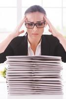 C'est trop. jeune femme déprimée en costume cravate regardant la pile de documents et tenant la tête dans les mains tout en étant assise sur son lieu de travail photo