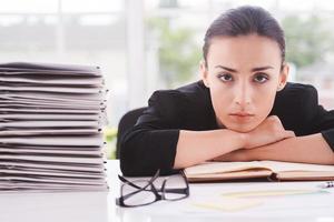 cadre surmené. jeune femme déprimée en tenues de soirée regardant la caméra et penchant son visage à la table avec une pile de documents près de photo