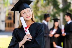 rêvant d'un avenir radieux. jeune femme réfléchie en robes de graduation tenant un diplôme et regardant loin tandis que ses amis se tenant en arrière-plan photo