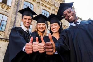 confiants dans leur avenir fructueux. vue en angle bas de quatre diplômés universitaires en robes de graduation debout près l'un de l'autre et montrant leurs pouces vers le haut photo