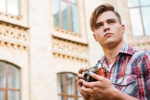 photographe inspiré. vue en angle bas d'un beau jeune homme tenant un appareil photo vintage et regardant loin tout en se tenant à l'extérieur