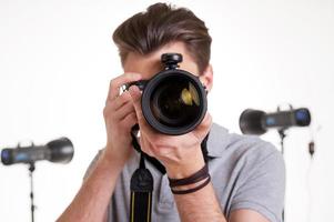 sourire à la caméra jeune homme en polo vous tirant avec un appareil photo numérique tout en se tenant en studio avec un équipement d'éclairage sur fond