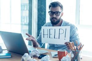 demander de l'aide. jeune homme barbu frustré tenant un morceau de papier et demandant de l'aide alors qu'il était assis à son lieu de travail au bureau photo