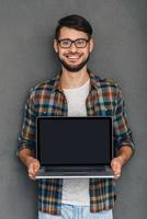 faites attention à ce jeune homme confiant montrant son ordinateur portable et regardant la caméra avec le sourire en se tenant debout sur fond gris photo