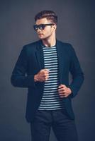 tendance et stylé. confiant jeune bel homme à lunettes de soleil ajustant sa veste et regardant ailleurs en se tenant debout sur fond gris photo