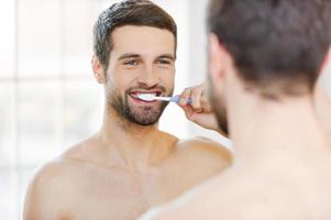 se brosser les dents le matin. vue arrière du beau jeune homme barbe se brosser les dents et souriant debout contre un miroir photo