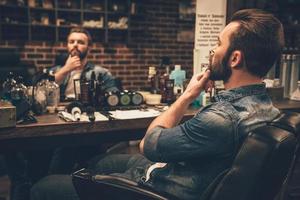 bonne mine. vue latérale d'un beau jeune homme barbu regardant son reflet dans le miroir et gardant la main sur le menton tout en étant assis sur une chaise au salon de coiffure photo