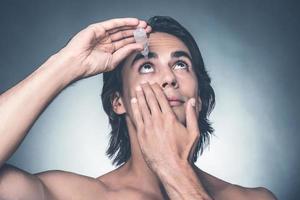 en prenant bien soin de ses yeux. portrait de jeune homme torse nu mettant des gouttes dans les yeux en se tenant debout sur fond gris photo