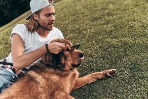 chien avec style. beau jeune homme mettant des lunettes de soleil sur son chien allongé sur l'herbe dans le parc photo