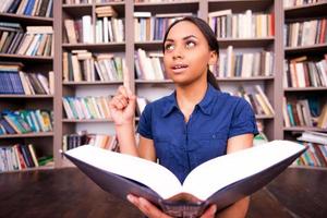 juste inspiré. Étudiante africaine surprise tenant un livre et pointant vers le haut alors qu'elle était assise par terre dans la bibliothèque photo