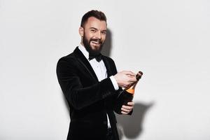 petit parti n'a jamais tué personne. beau jeune homme en costume complet ouvrant une bouteille de champagne et souriant en se tenant debout sur fond gris photo