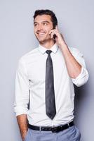 bonne conversation d'affaires. beau jeune homme en chemise et cravate parlant au téléphone mobile et souriant en se tenant debout sur fond gris photo