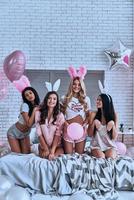 s'amuser. quatre jeunes femmes souriantes ludiques dans des oreilles de lapin faisant une grimace et gesticulant assis sur le lit avec des ballons autour photo