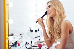 ajouter de la couleur à mes joues. vue latérale d'une jeune femme joyeuse qui se maquille et regarde son reflet dans le miroir tout en étant assise à la coiffeuse photo