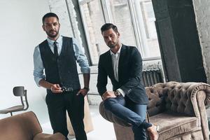 confiance et style. deux beaux jeunes hommes en costume regardant la caméra tout en se reposant à l'intérieur photo