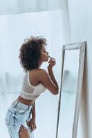 elle fera fondre votre coeur. séduisante jeune femme africaine mettant du rouge à lèvres debout devant le miroir à la maison photo