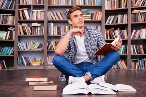 préparer ses examens finaux. jeune homme réfléchi tenant un bloc-notes et touchant son menton avec un stylo assis contre une étagère photo