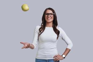 belle jeune femme jetant une pomme et souriant debout sur fond gris photo