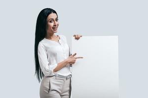 placez de nouvelles idées ici jeune femme confiante en chemise blanche pointant sur un tableau vierge en se tenant debout sur fond gris photo