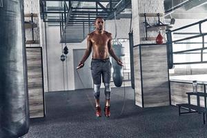 formation pour devenir le meilleur. toute la longueur du jeune homme africain en vêtements de sport sautant à la corde tout en faisant de l'exercice dans la salle de sport photo