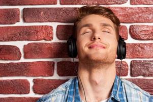 appréciant sa musique préférée. beau jeune homme écoutant le lecteur mp3 et gardant les yeux fermés tout en se tenant contre le mur de briques photo