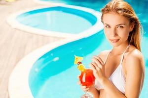 juste moi et mon cocktail d'été. belle jeune femme en bikini blanc tenant un cocktail et regardant la caméra tout en se relaxant au bord de la piscine photo