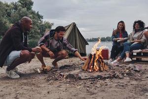 tout simplement relaxant. groupe de jeunes en tenue décontractée souriant tout en profitant d'une fête sur la plage près du feu de camp photo