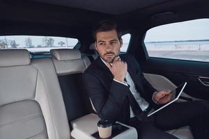 jeune perfectionniste. beau jeune homme en costume complet travaillant à l'aide d'une tablette numérique assis dans la voiture photo