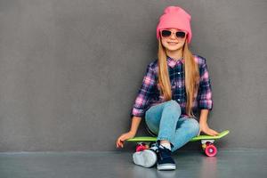 mignonne insouciante avec planche à roulettes. pleine longueur de joyeuse petite fille à lunettes de soleil regardant la caméra avec le sourire tout en étant assis sur une planche à roulettes sur fond gris photo