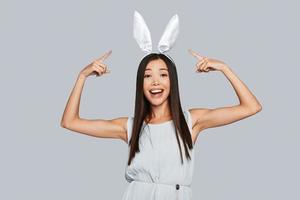 regardez belle jeune femme asiatique pointant sur ses oreilles de lapin et souriant debout sur fond gris photo