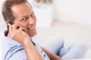 bonne nouvelle des amis. vue de dessus d'un homme mûr joyeux parlant au téléphone portable et regardant par-dessus l'épaule tout en étant assis sur le canapé à la maison photo