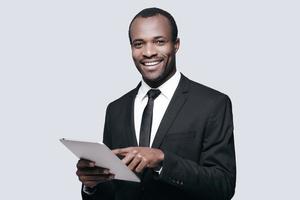 se sentir heureux au travail. beau jeune homme africain souriant et utilisant une tablette numérique en se tenant debout sur fond gris photo