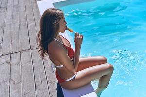 C'est délicieux. belle jeune femme en maillot de bain mangeant des glaces assise au bord de la piscine en plein air photo