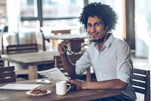 bonjour jeune homme africain tenant un journal et regardant la caméra avec le sourire assis au café