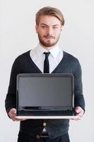 copier l'espace sur le moniteur. beau jeune homme tenant un ordinateur portable et souriant en se tenant debout sur fond gris photo